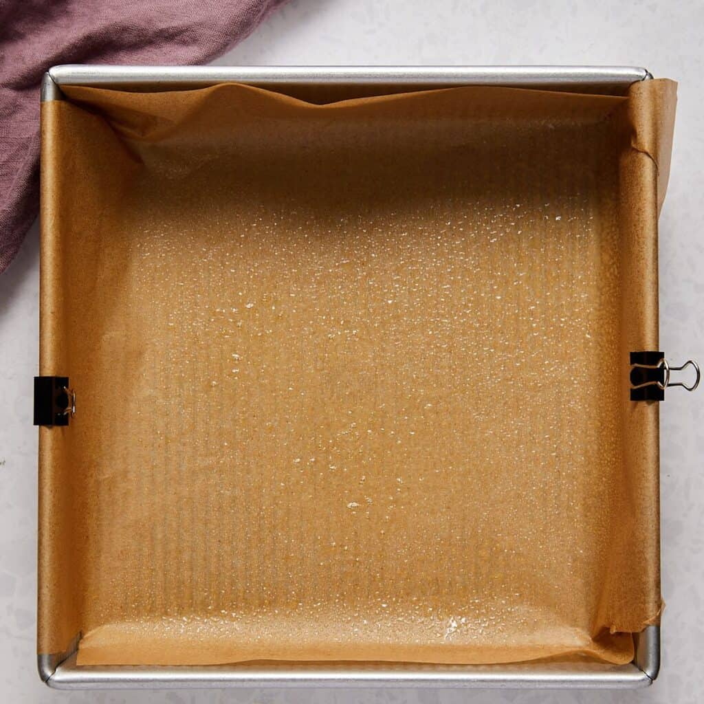 A square baking pan lined with brown parchment paper, secured at the corners with metal binder clips and lightly sprayed with oil, sits on a light surface next to a folded mauve cloth.