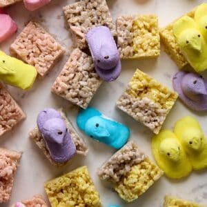 Colorful marshmallow chick candies and rice crispy treats are arranged on a light marble surface. The chicks are pink, purple, blue, and yellow, scattered among the square treats.