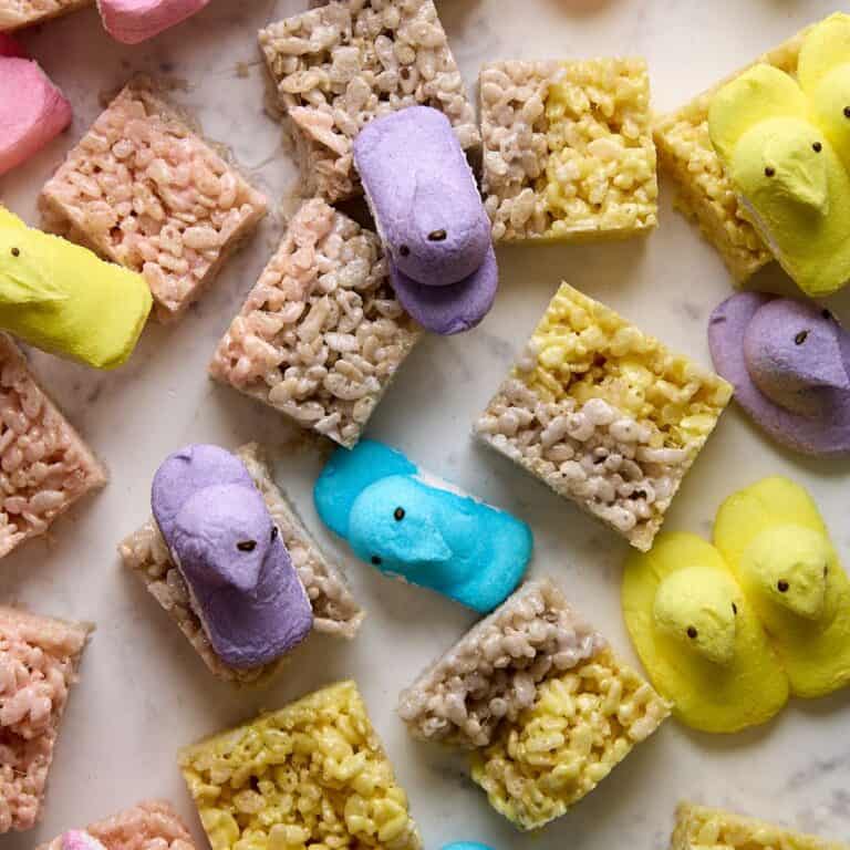 Colorful marshmallow chick candies and rice crispy treats are arranged on a light marble surface. The chicks are pink, purple, blue, and yellow, scattered among the square treats.