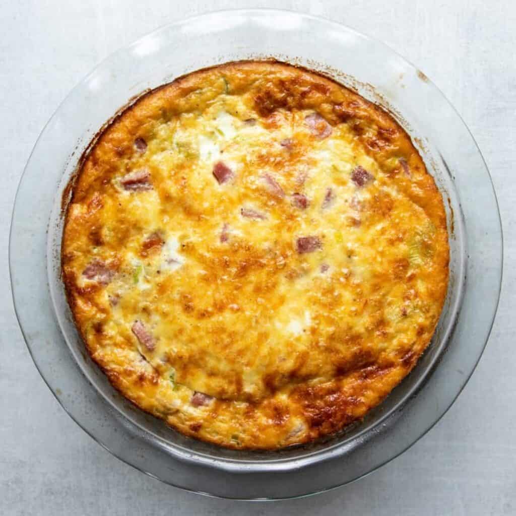 A baked crustless leek quiche in a glass pie dish, golden on top with visible ham and Gruyère.
