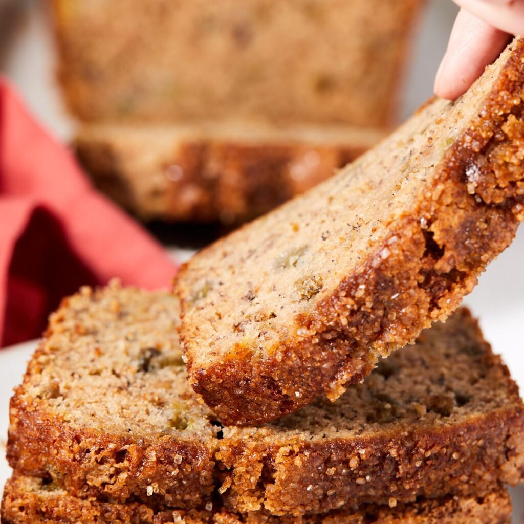 A close-up of a hand lifting a slice of moist banana bread from a stack, with a red cloth and a blurred loaf in the background. The bread has a golden-brown, crumbly crust and visible bits of banana and nuts.