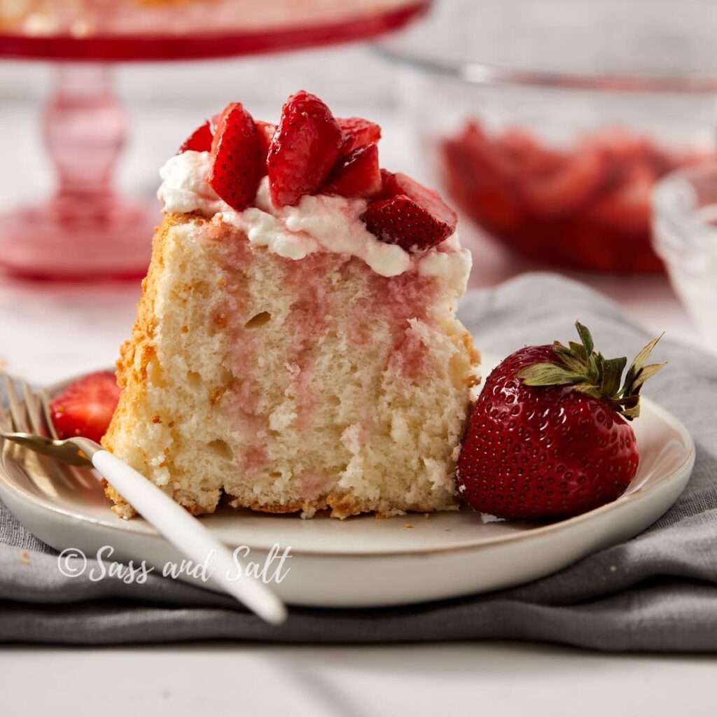 A slice of strawberry shortcake topped with whipped cream and fresh strawberries sits on a plate with a fork and whole strawberry, with more strawberries and cake in the background.
