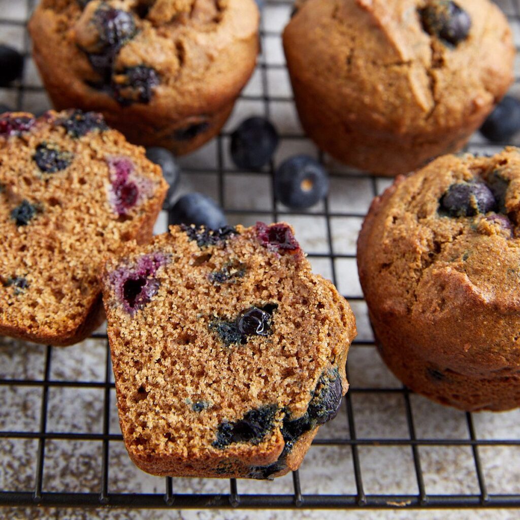 Blueberry muffins cooling on a wire rack, with one muffin cut in half to show its moist, blueberry-filled interior. Whole blueberries are scattered nearby.