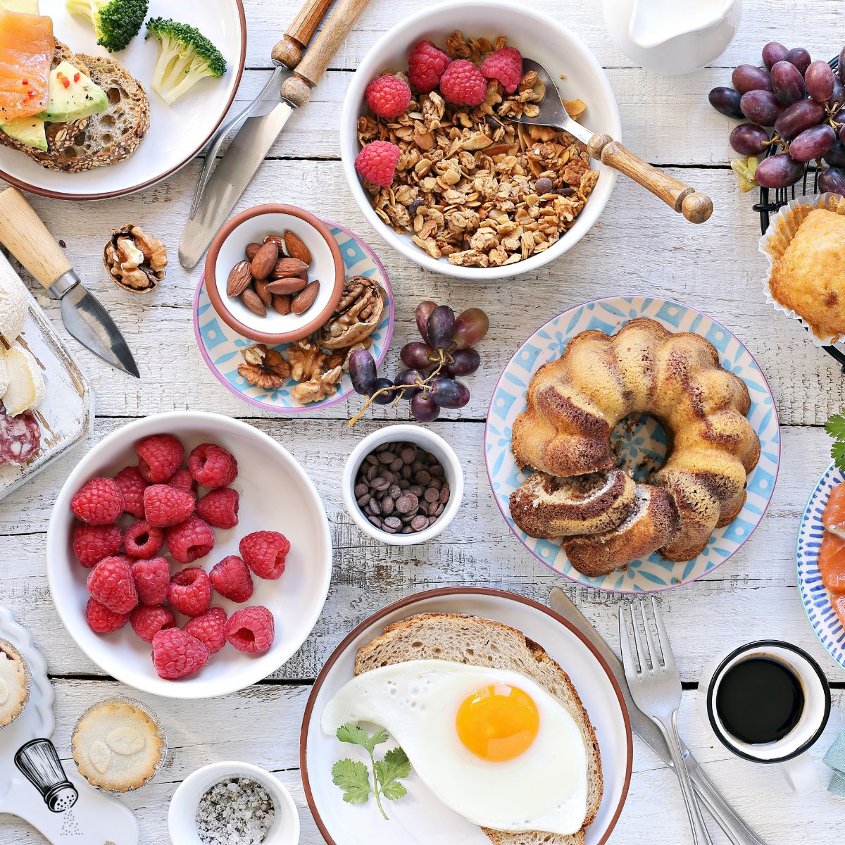 A breakfast spread on a white wooden table, featuring a fried egg on toast, raspberries, grapes, bundt cake, granola with raspberries, nuts, chocolate chips, coffee, and various cheeses and vegetables.