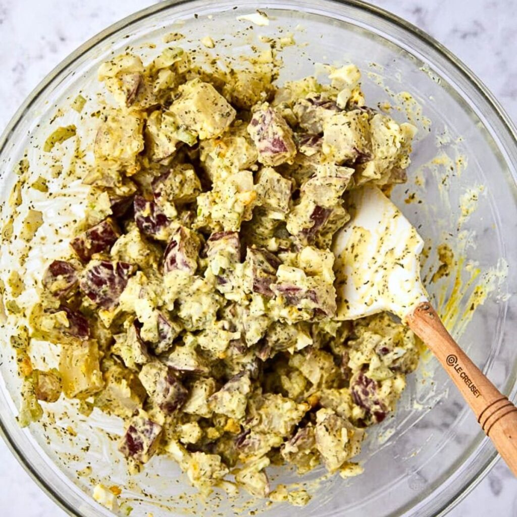 A glass bowl filled with creamy potato salad mixed with herbs, with a white spatula resting on the side. The salad features chunks of red-skinned potatoes coated in dressing.