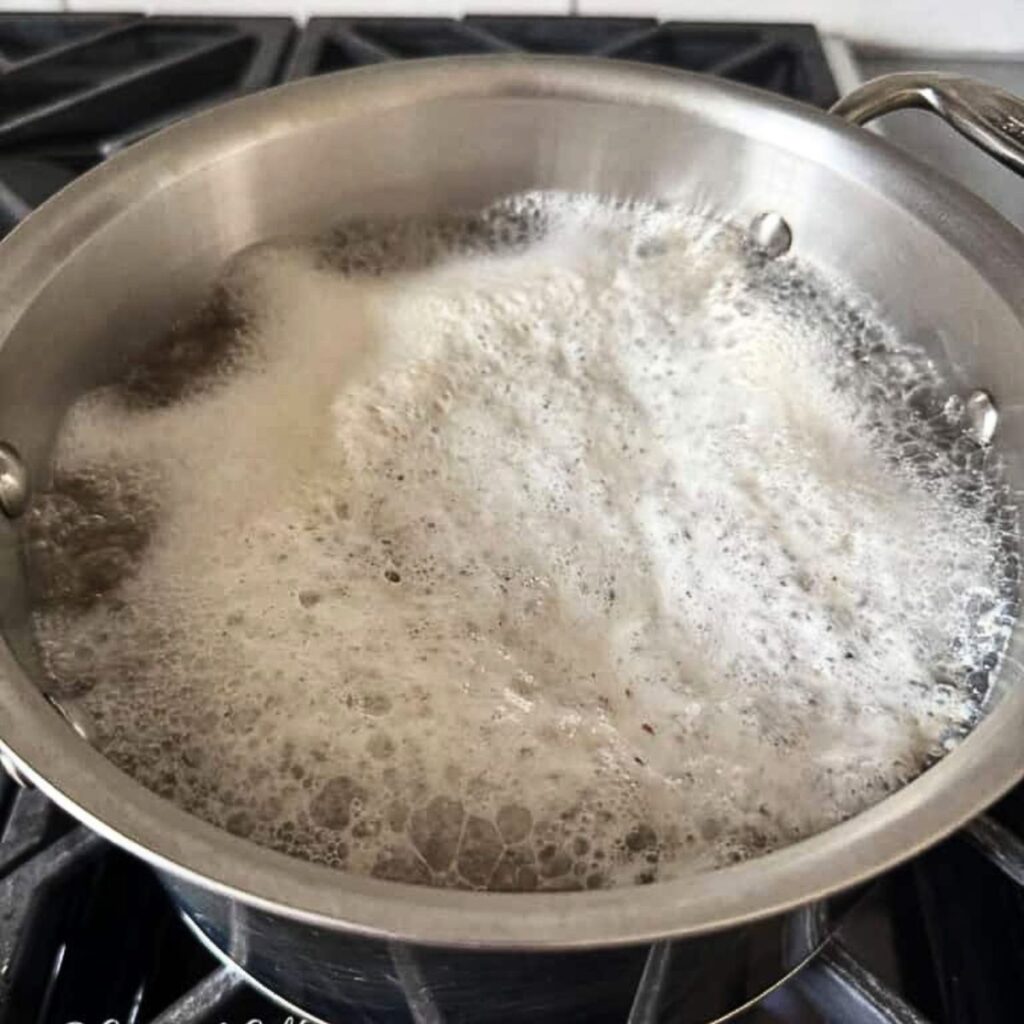 A stainless steel pot filled with foamy, bubbling water sits on a stove with red potatoes in pot.