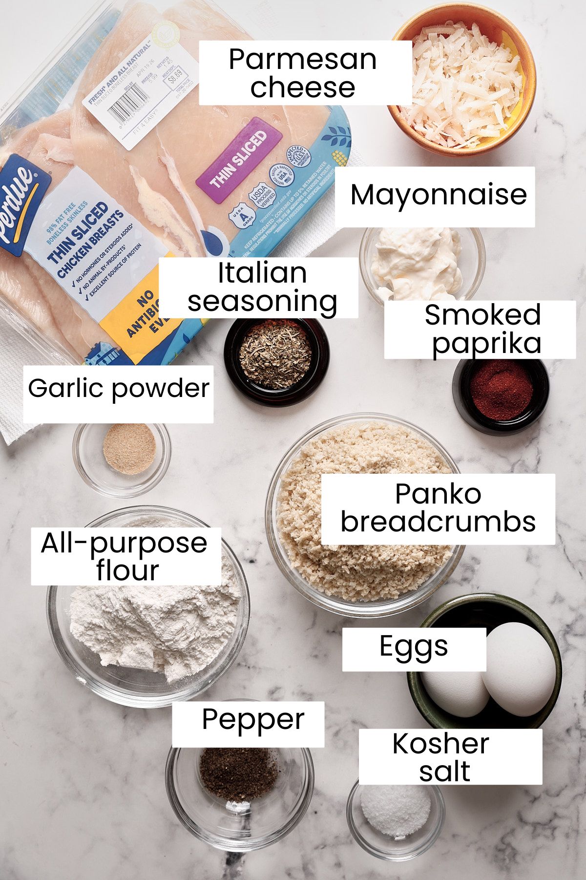 An overhead view of ingredients for crispy air fryer chicken cutlets in bowls and plates labeled: thin sliced chicken breasts, Parmesan cheese, mayonnaise, Italian seasoning, smoked paprika, garlic powder, panko breadcrumbs, flour, eggs, pepper, and kosher salt.