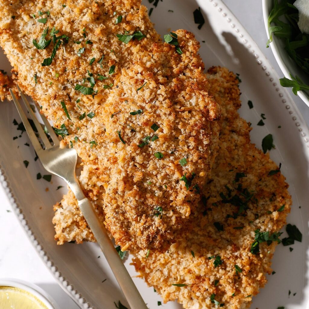 A plate with several pieces of golden, crispy chicken cutlets air fryer garnished with chopped parsley, accompanied by a gold fork. The dish is set on a white plate, with part of a lemon and greens visible nearby.