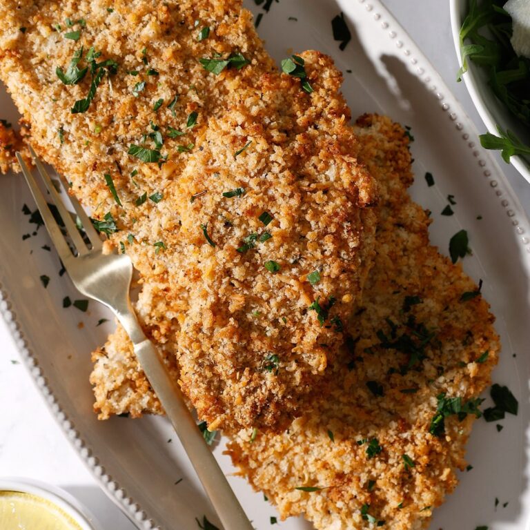 A plate with several pieces of golden, crispy chicken cutlets air fryer garnished with chopped parsley, accompanied by a gold fork. The dish is set on a white plate, with part of a lemon and greens visible nearby.