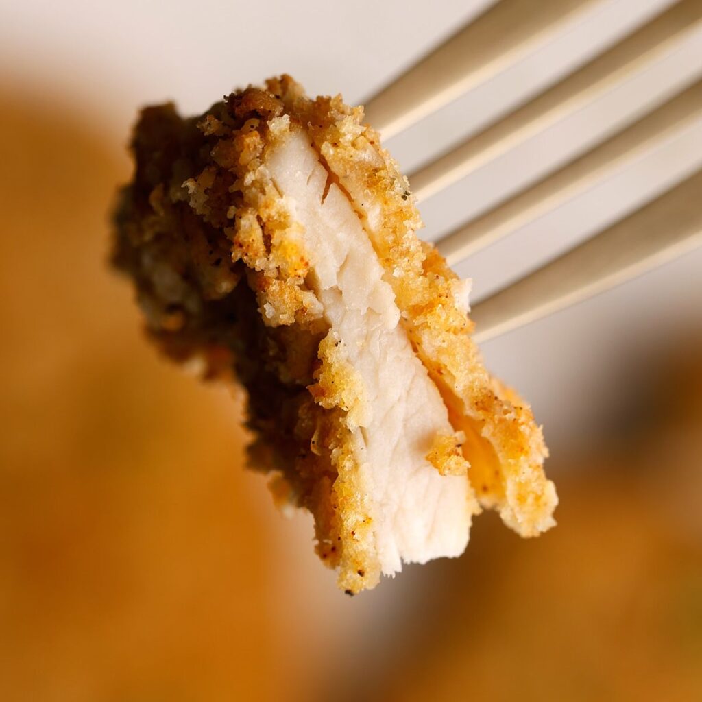  A close-up of a fork holding a piece of crispy air fryer thin sliced chicken breast, showing the golden-brown panko crust and juicy white meat inside.