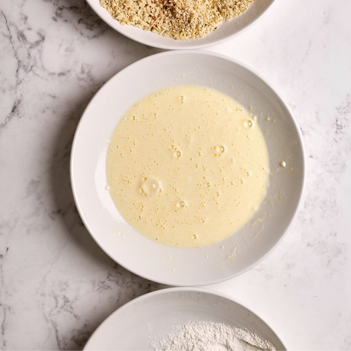 A white bowl of beaten eggs surrounded by plates of flour and panko breadcrumbs on a marble surface, the breading station for air fryer thin sliced chicken breast.