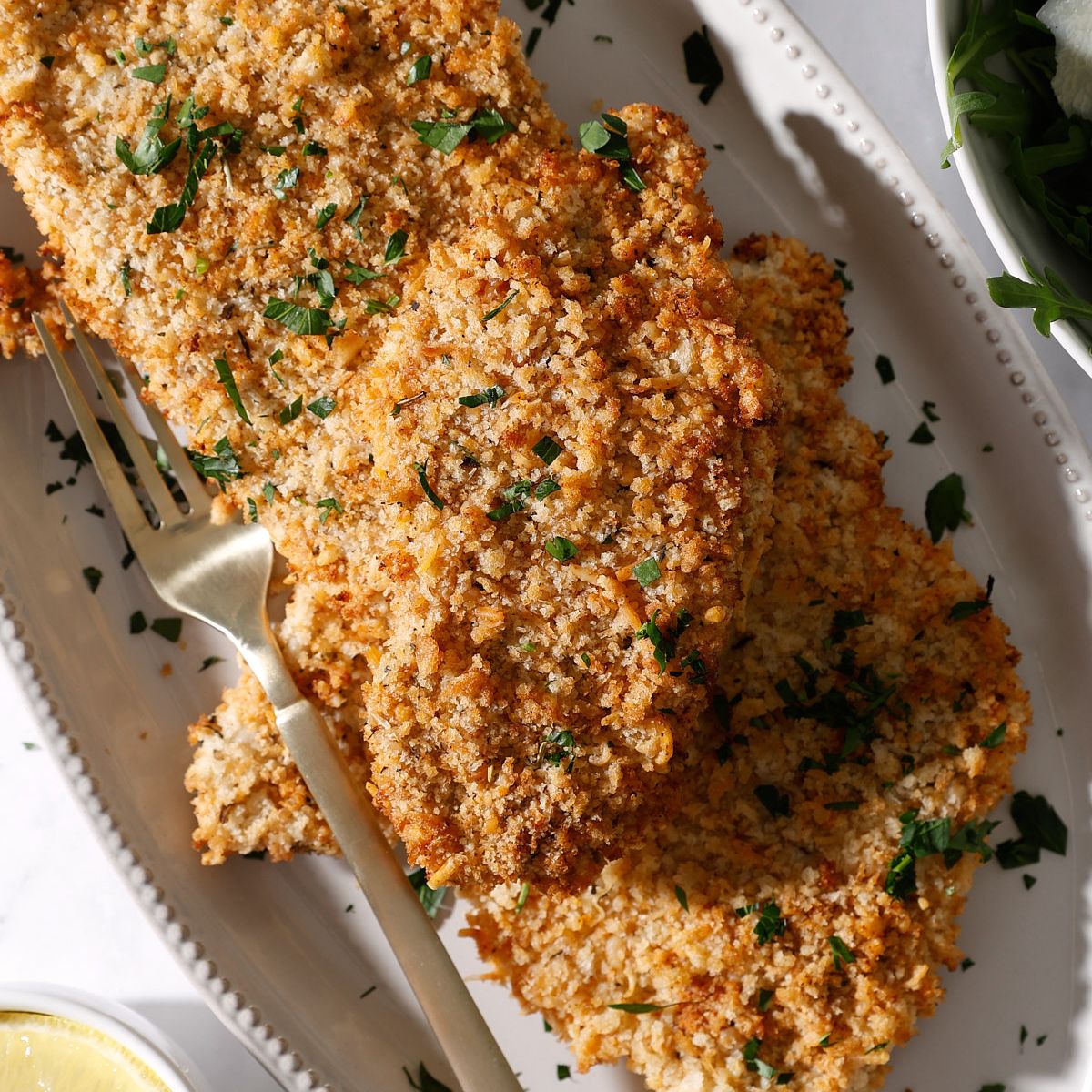 A plate with several pieces of golden, crispy chicken cutlets air fryer garnished with chopped parsley, accompanied by a gold fork. The dish is set on a white plate, with part of a lemon and greens visible nearby.