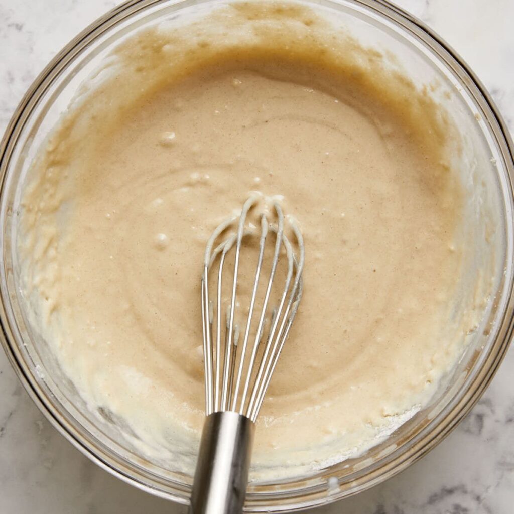 A metal whisk resting in a glass bowl filled with smooth, light tan batter on a white marble surface.