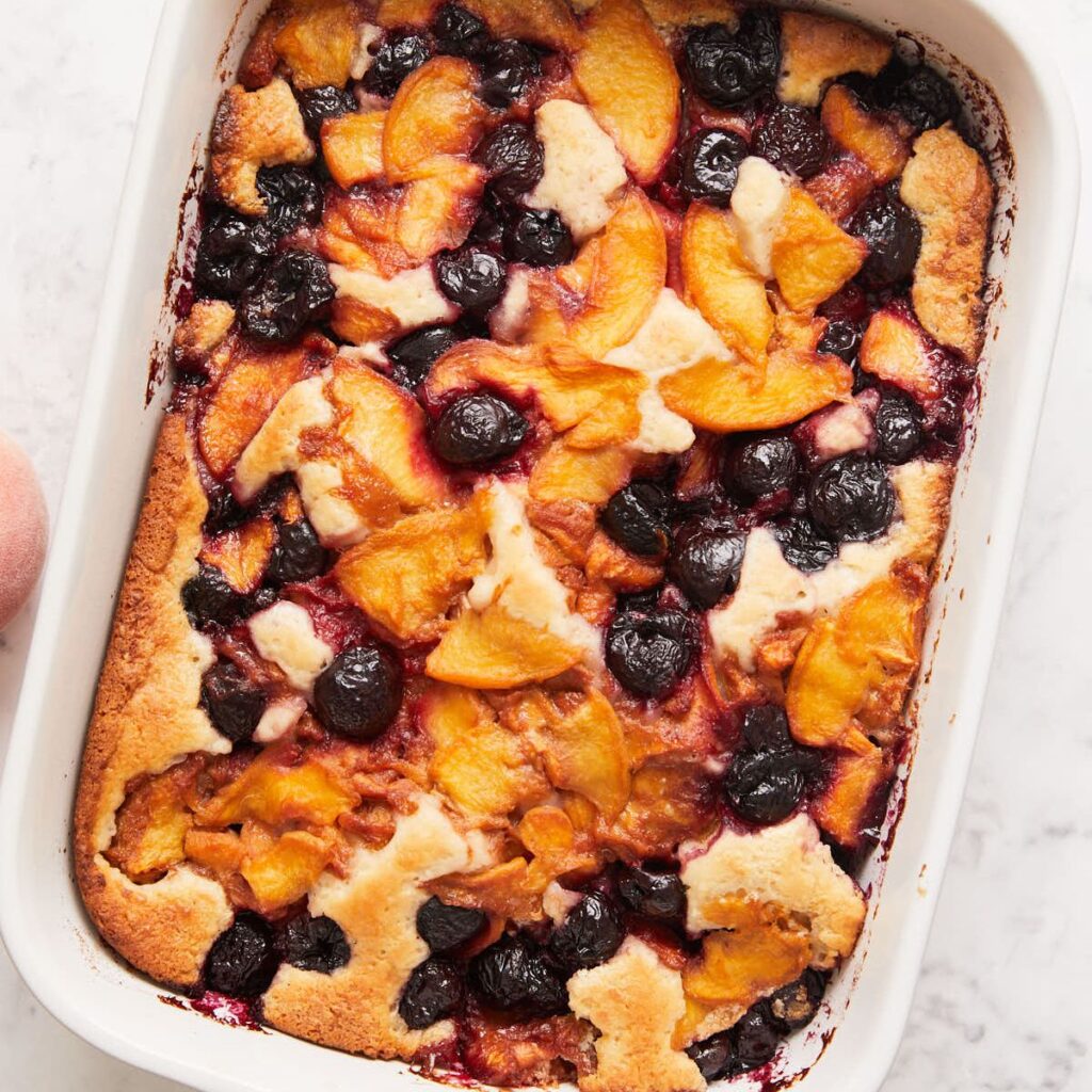 A baked fruit cobbler in a white dish, featuring golden peach slices and dark purple berries on a lightly browned, cake-like crust.