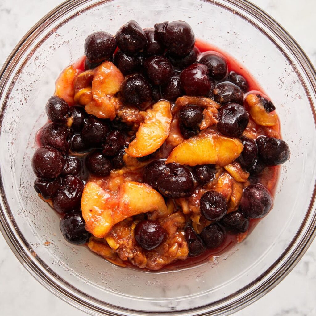 A clear glass bowl filled with a mixture of dark cherries and peach slices in a syrupy liquid, sitting on a white marble surface.