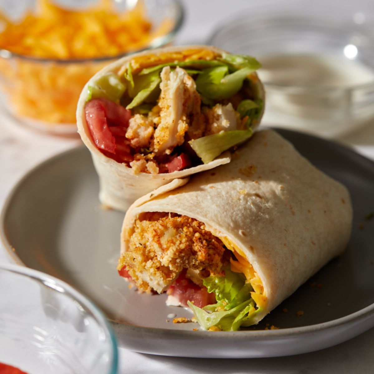 A crispy chicken wrap cut in half, filled with breaded chicken, lettuce, tomatoes, and shredded cheese, served on a gray plate with bowls of cheese and ranch dressing in the background.