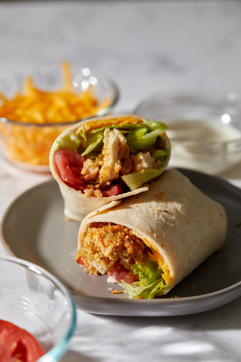 A halved crispy chicken wrap on a gray plate with the cross-section facing the camera, showing layers of breaded chicken, cheese, lettuce, and tomato.