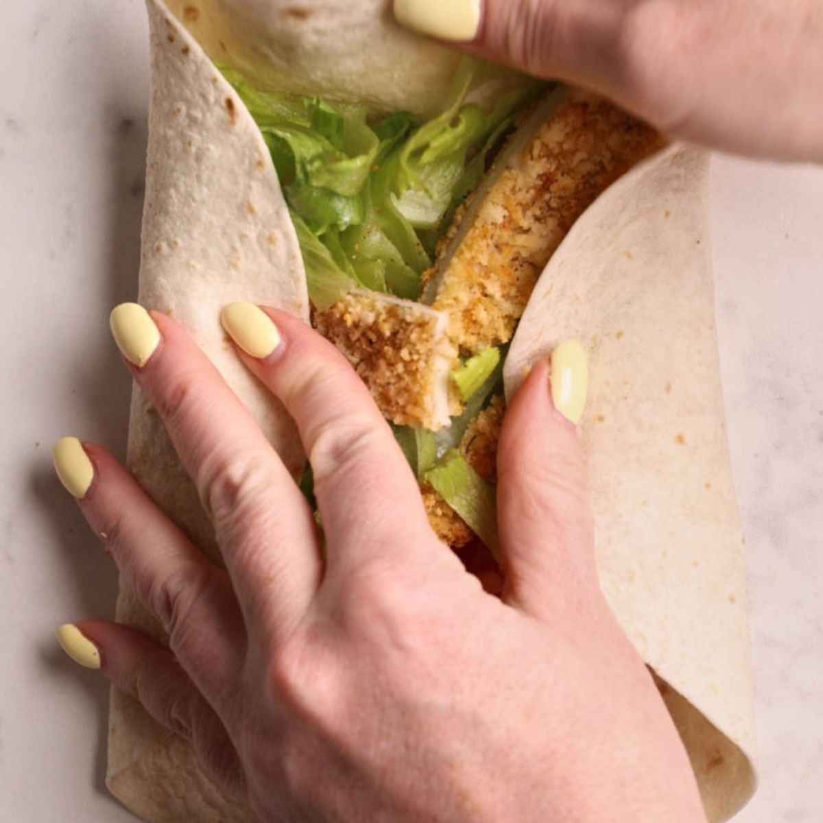 Hands folding a loaded tortilla into a crispy chicken wrap, rolling from the bottom up to seal the seam.