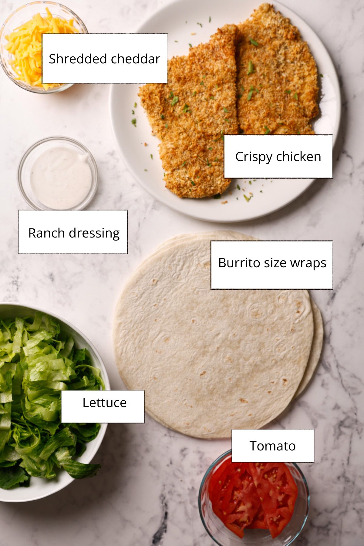 Crispy chicken wrap ingredients labeled on a white surface: shredded cheddar, sliced crispy chicken cutlets, ranch dressing, flour tortillas, shredded romaine, and sliced tomatoes.