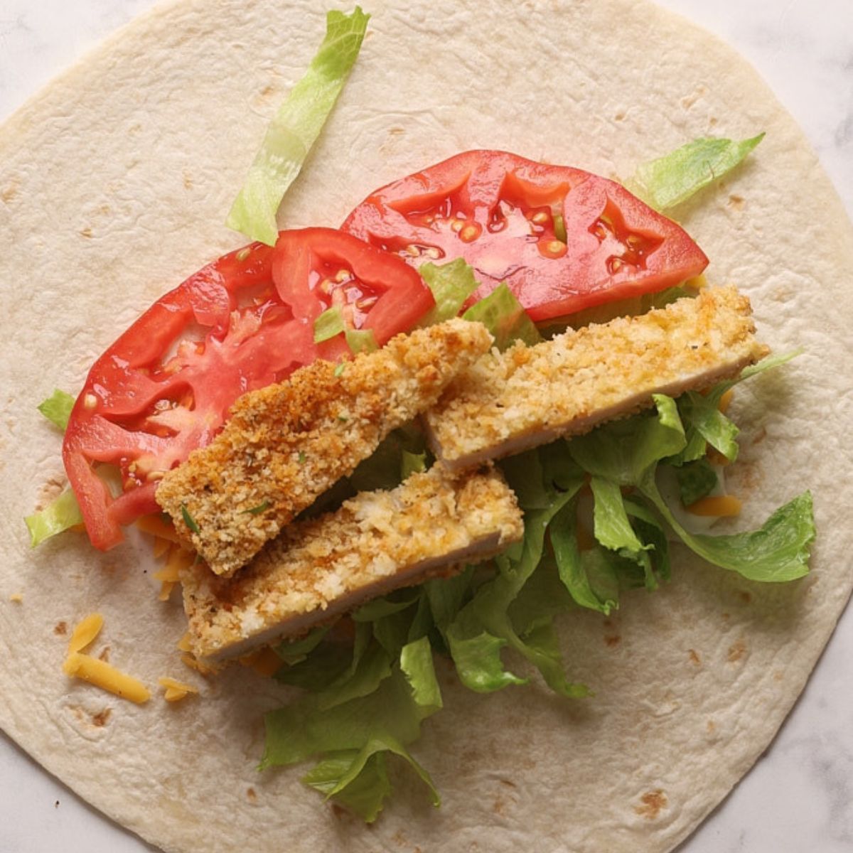 A flour tortilla layered with crispy chicken strips, shredded cheddar, romaine lettuce, and tomato slices, ready to be rolled into a wrap.