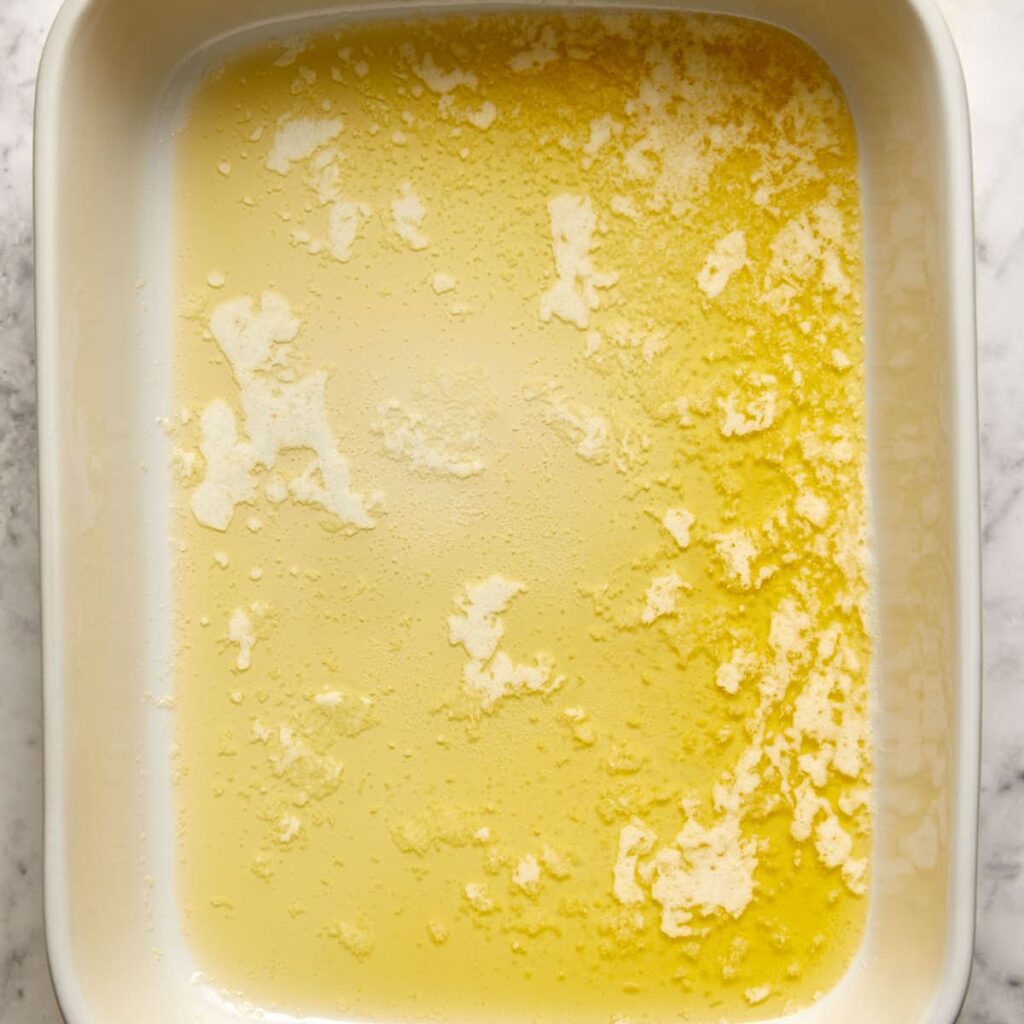 White rectangular baking dish with melted butter spread unevenly across the bottom, viewed from above on a light-colored surface.