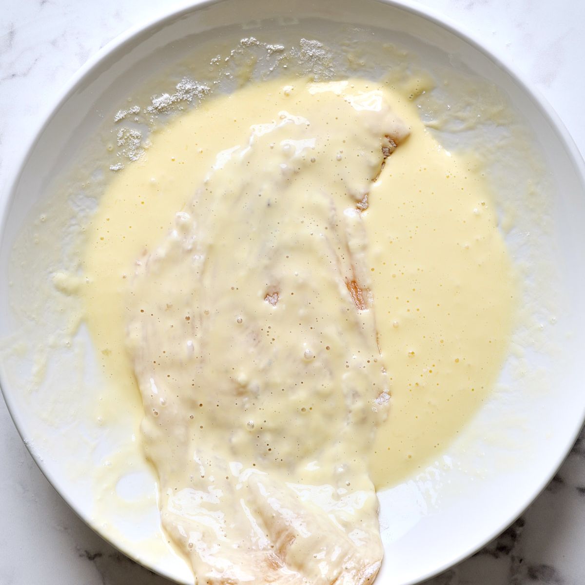 A thin sliced chicken breast coated in a pale yellow egg-mayo wash in a white bowl, the second step of breading for the air fryer.