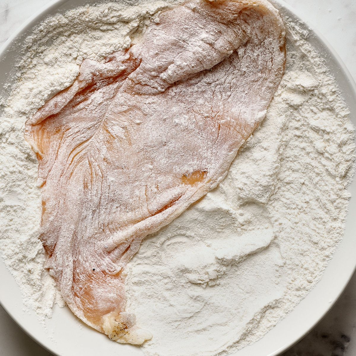 A raw thin sliced chicken breast coated with flour on a large white plate, the first dredging step for air fryer chicken cutlets.