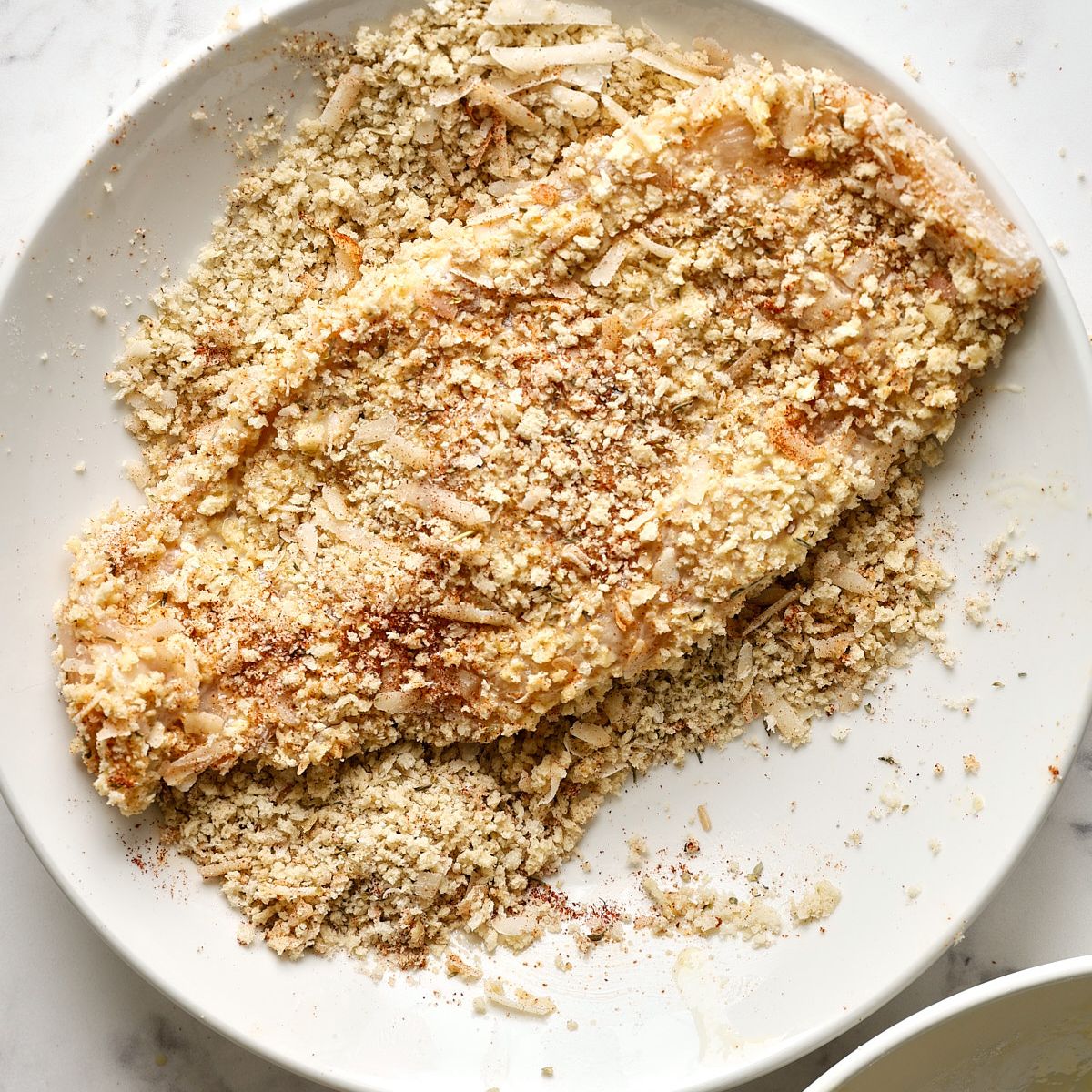 A raw thin sliced chicken breast pressed into a panko-Parmesan breading mixture on a white plate, ready for the air fryer.