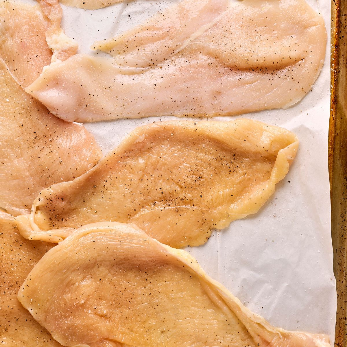 Raw, thinly sliced chicken breasts seasoned with salt and pepper, arranged in a single layer on a sheet of white parchment paper, ready for the air fryer.