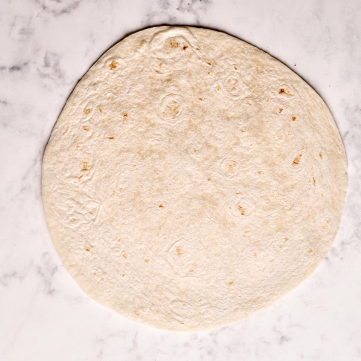 A warmed 10-inch flour tortilla flat on a marble surface, ready to be loaded with crispy chicken.