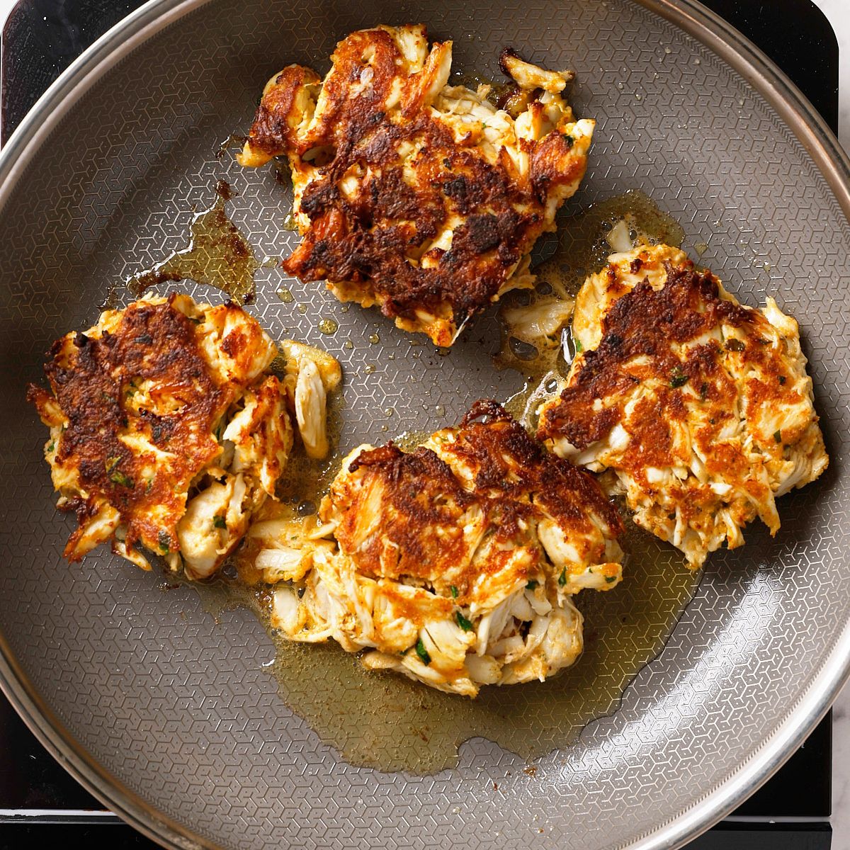 Four golden-brown crab cakes frying in oil on a nonstick skillet, with crispy edges and visible chunks of crab meat.