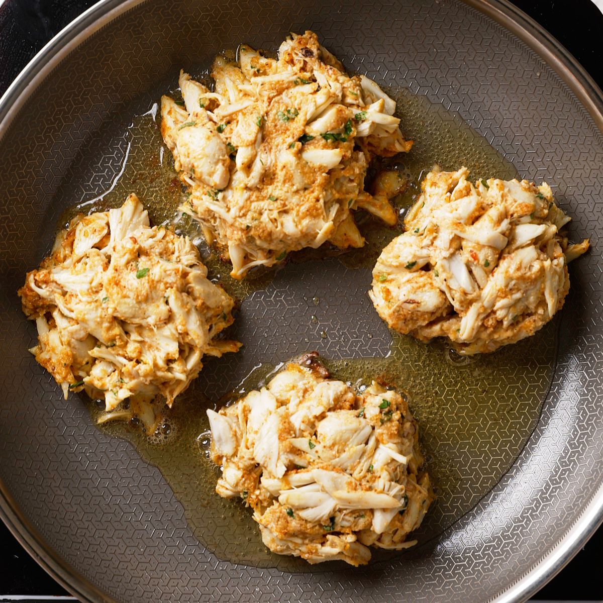 Four crab cakes cooking in a nonstick skillet with a bit of oil, showing golden brown edges and chunks of crab meat throughout.