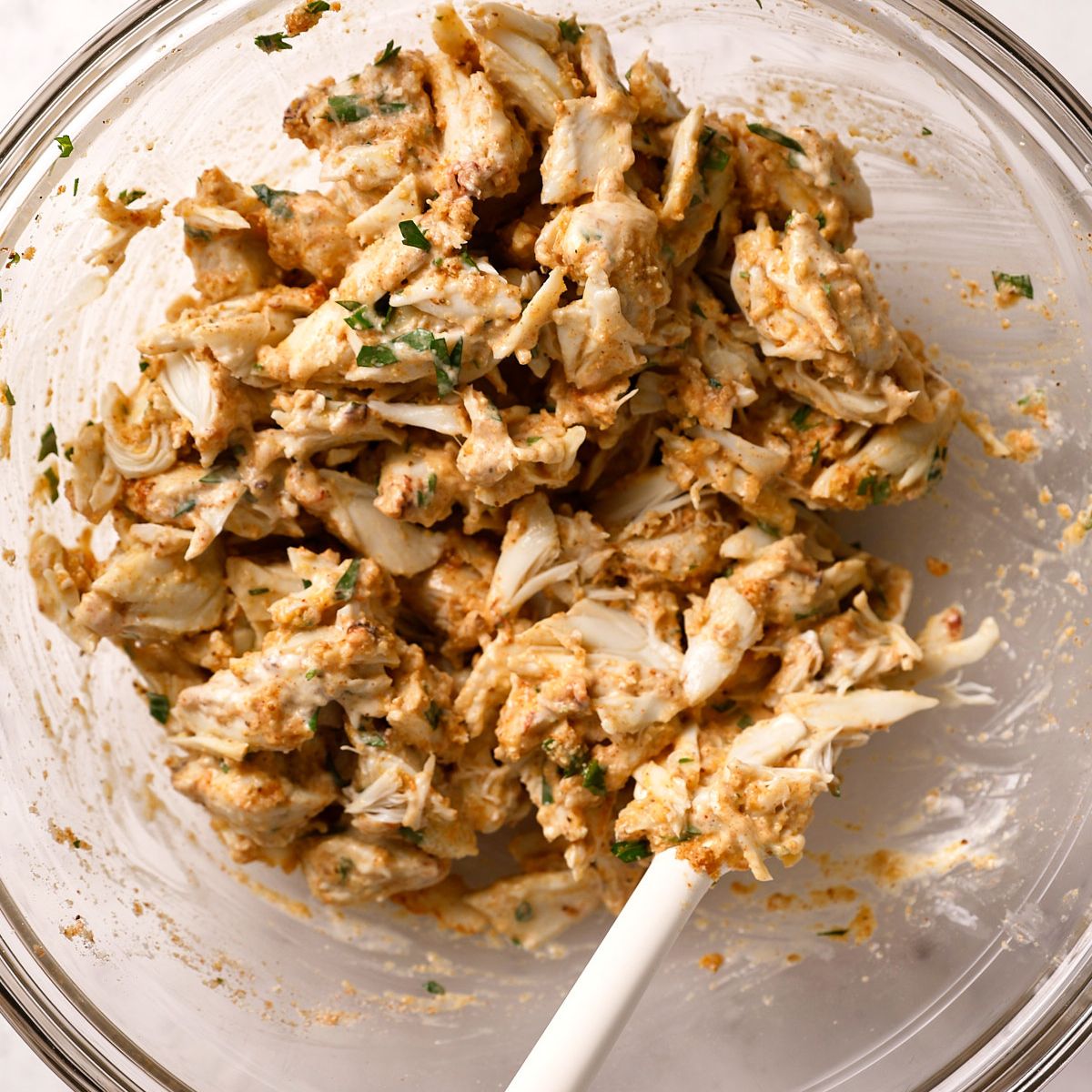 A glass bowl with a creamy, seasoned crab mixture being stirred by a white spatula; visible chunks of crab and herbs.