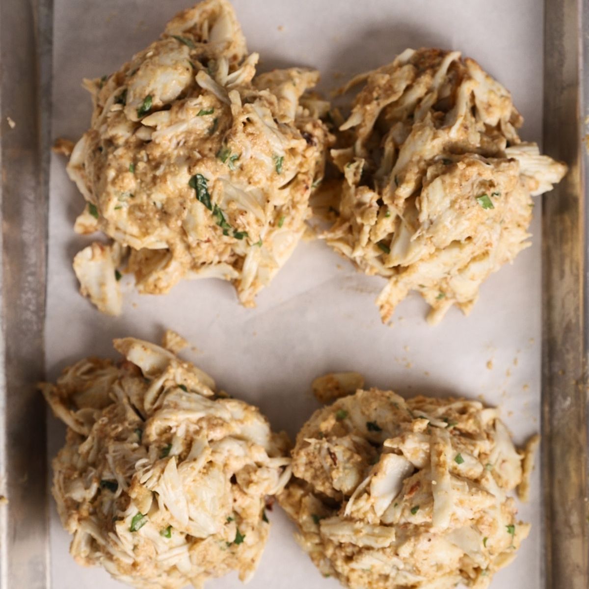 Four uncooked crab cakes, mixed with herbs and seasonings, are placed on a parchment-lined baking sheet.