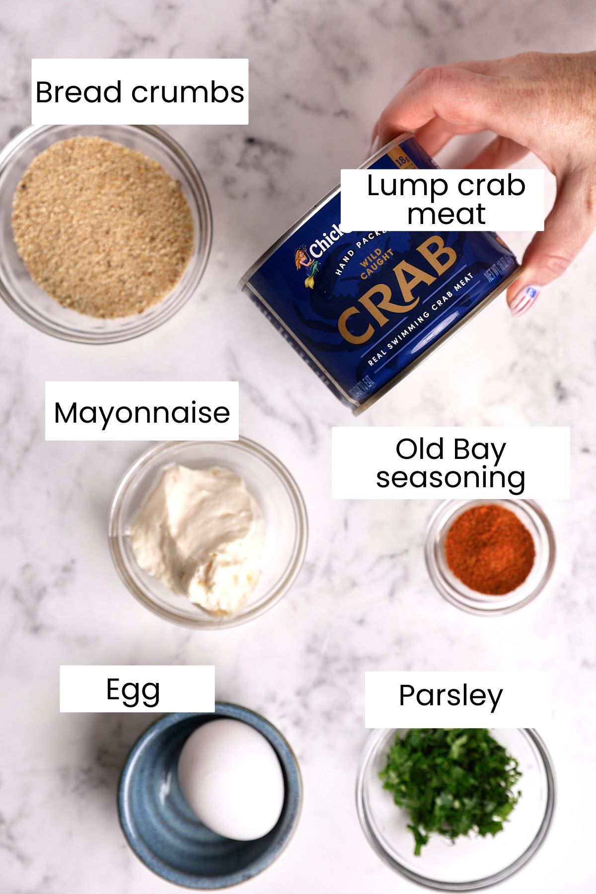 Six labeled ingredients on a marble surface: bread crumbs, lump crab meat, mayonnaise, Old Bay seasoning, an egg, and chopped parsley, each in separate bowls or containers.