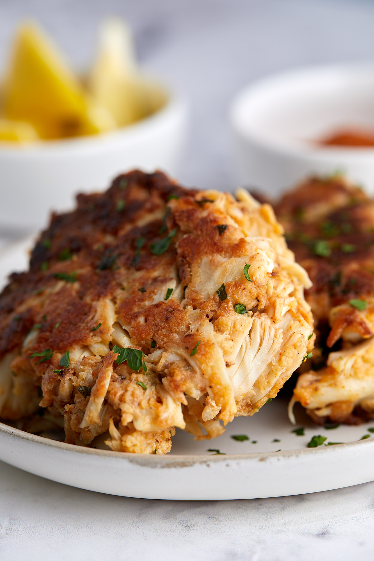 Two golden-brown crab cakes garnished with chopped herbs sit on a white plate. In the blurred background, there are lemon wedges and a small dish of dipping sauce.