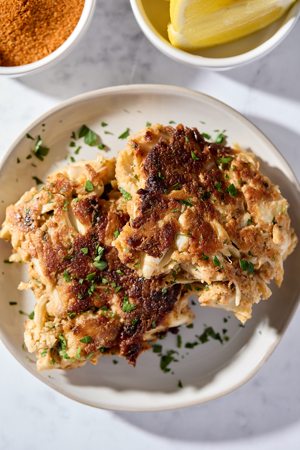 Two golden-brown crab cakes garnished with chopped parsley on a white plate, with a bowl of lemon wedges and a bowl of seasoning in the background.