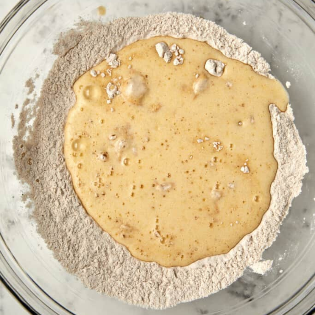 A glass bowl with dry flour mixture and wet ingredients just poured in, ready to be mixed for baking.