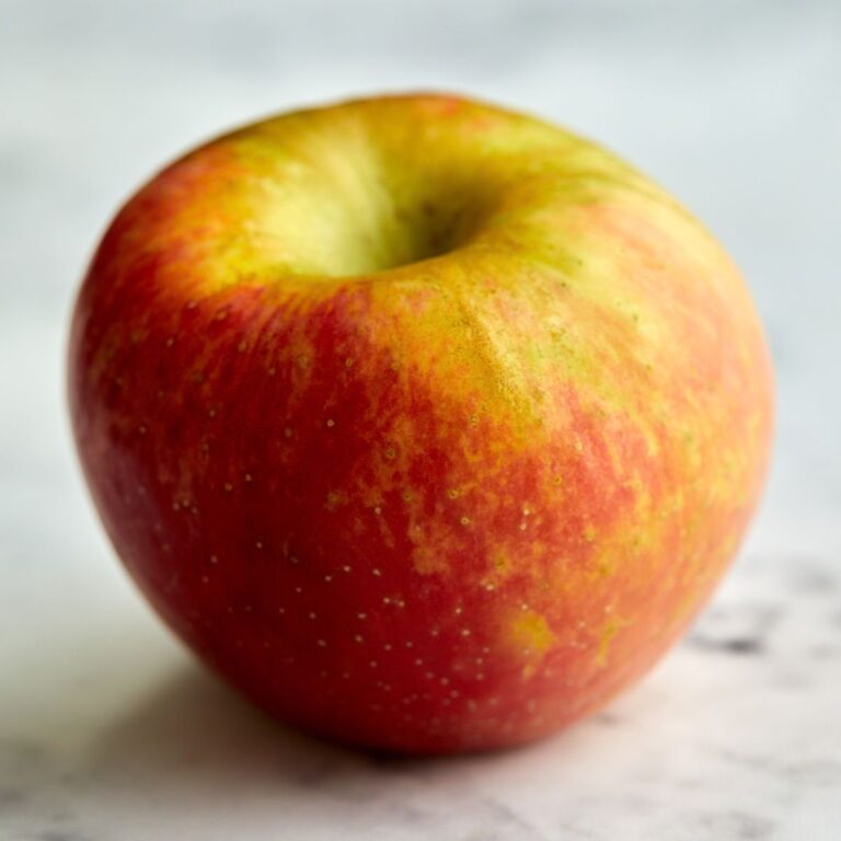 A close-up of a single red and yellow apple with a smooth surface, resting on a light, slightly blurred background.