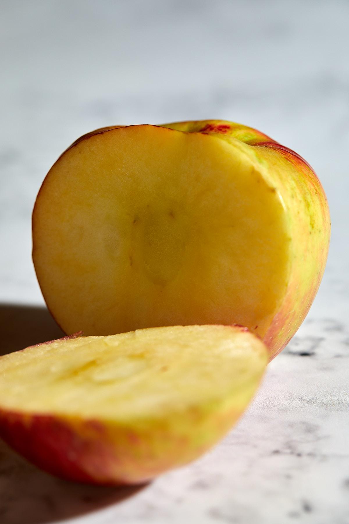 A close-up of an apple cut in half, showing its yellowish interior and red-green skin, placed on a light marble surface.