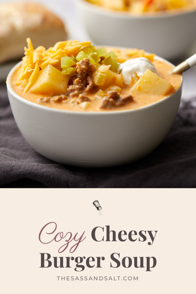 A bowl of cozy cheeseburger soup topped with shredded cheddar, pickles, and sour cream, with text below reading “Cozy Cheesy Burger Soup”.