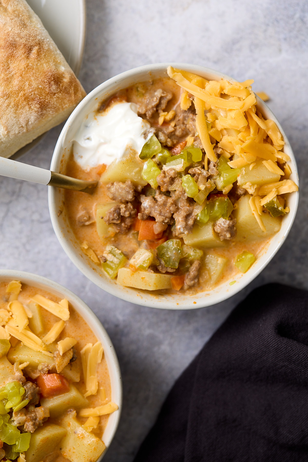 Two bowls of cheeseburger soup topped with shredded cheddar, sour cream, and diced dill pickles, served with a piece of crusty bread.
