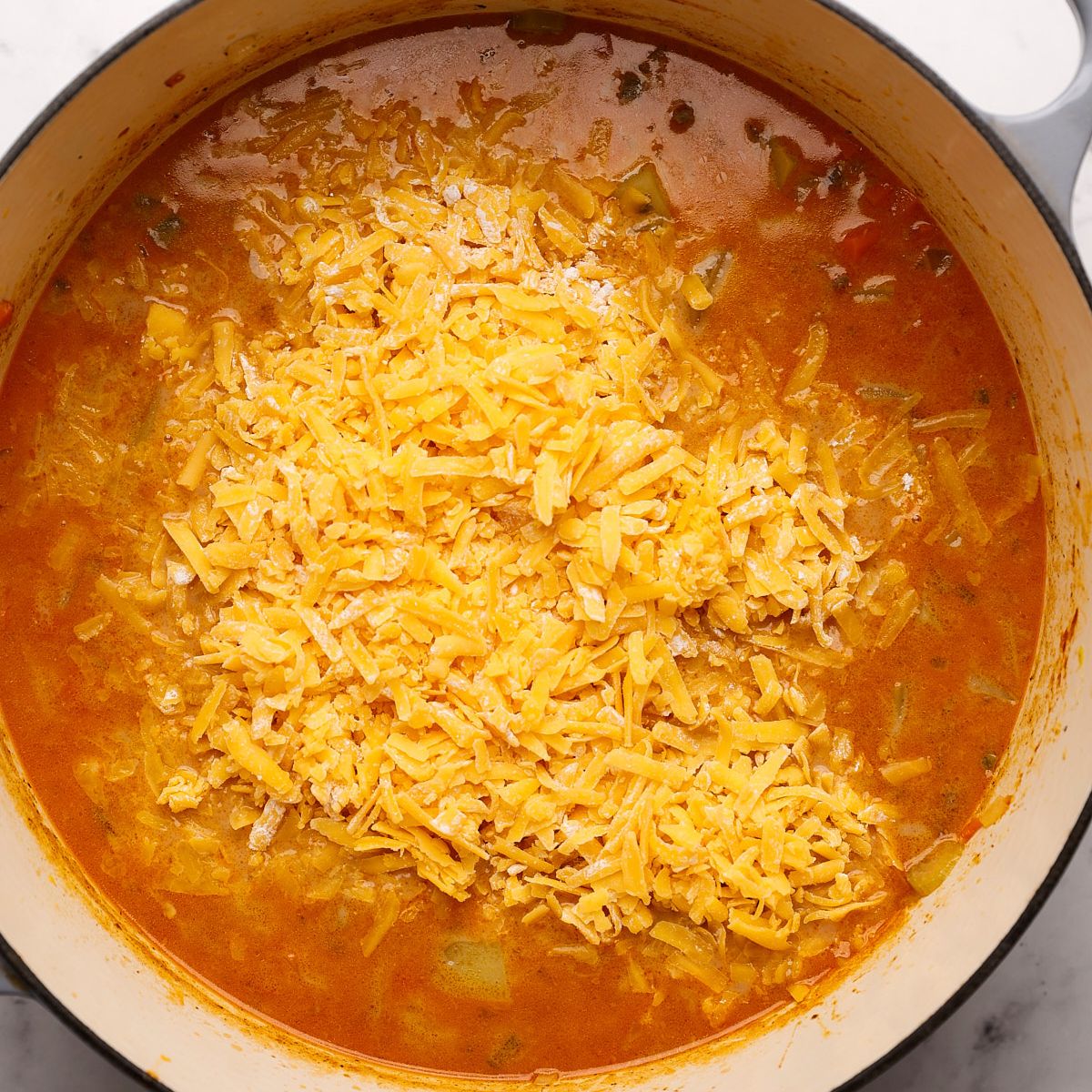 A mound of shredded cheddar cheese on top of cheeseburger soup in a Dutch oven, ready to be stirred in.