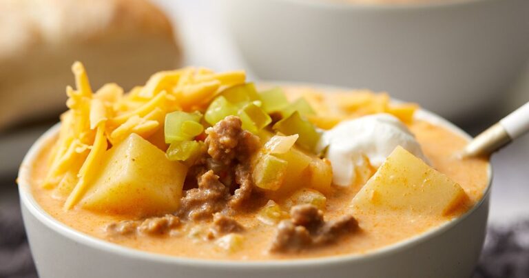 A bowl of cheeseburger potato soup topped with shredded cheddar, chopped pickles, and a dollop of sour cream.