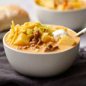 A bowl of creamy cheeseburger soup topped with shredded cheddar cheese, chopped pickles, and a dollop of sour cream.