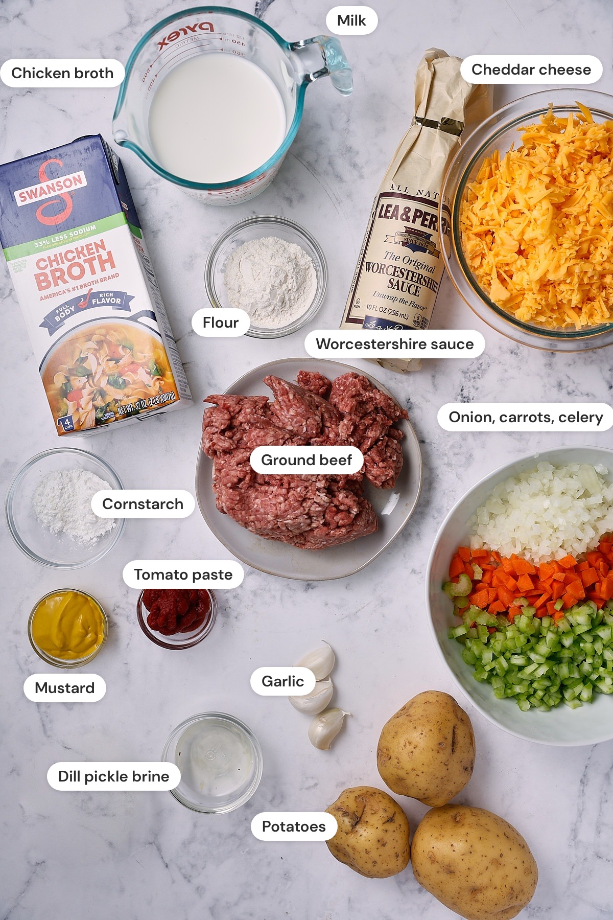 Ingredients for cheeseburger soup on a marble surface, including ground beef, Yukon Gold potatoes, diced onion, carrots, and celery, a block of sharp cheddar, chicken broth, milk, tomato paste, yellow mustard, Worcestershire sauce, garlic, flour, cornstarch, and dill pickle brine.
