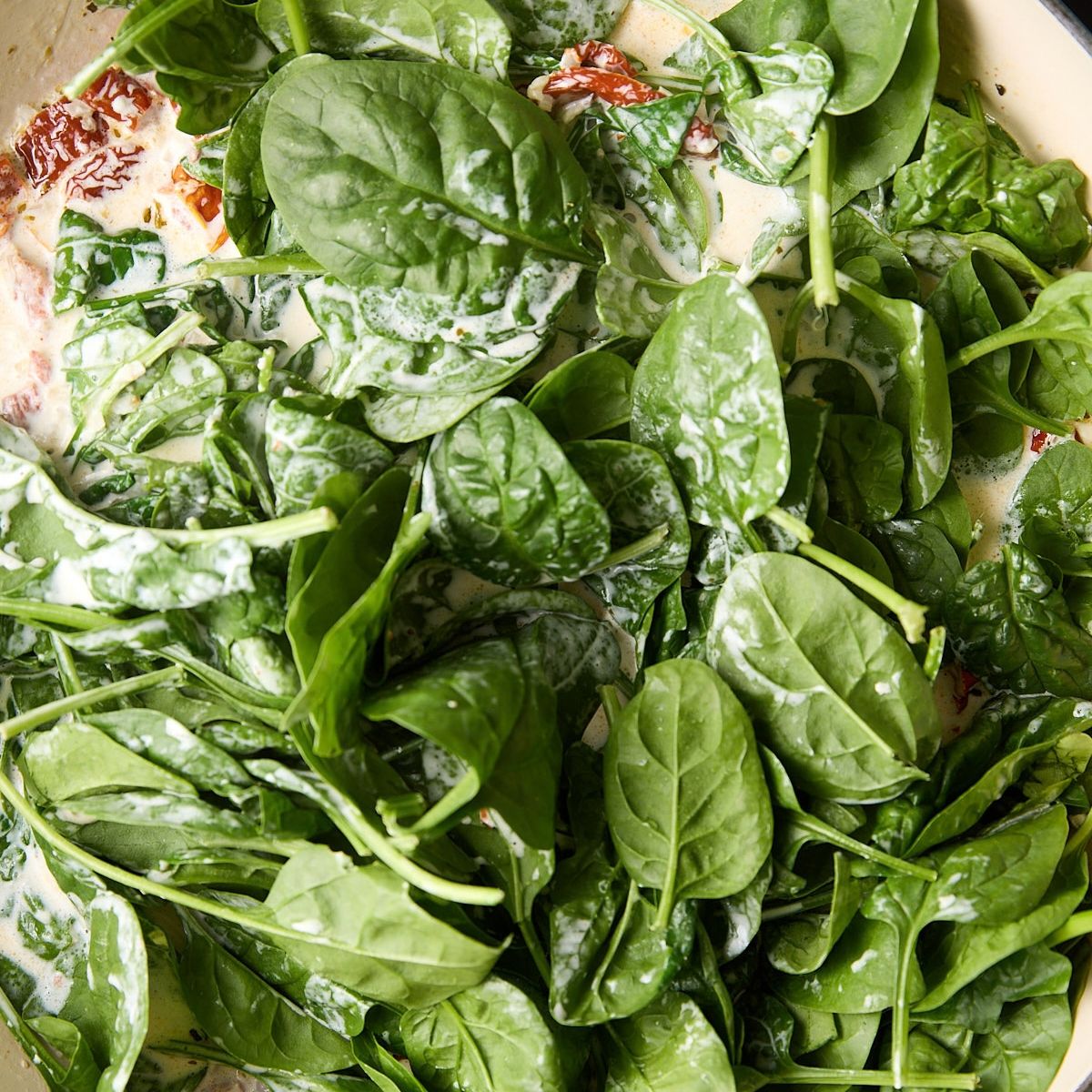 A mound of fresh baby spinach piled on top of cream sauce in a skillet, not yet wilted.