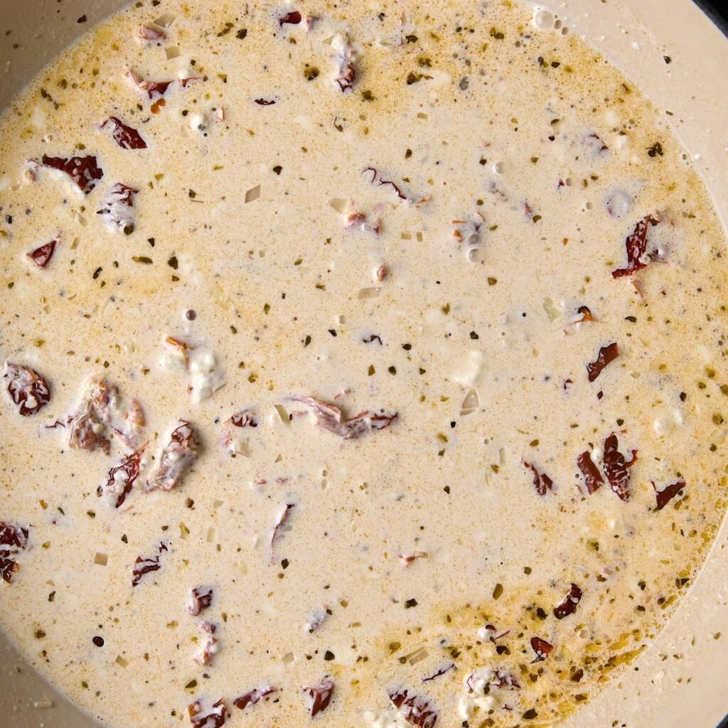 Pale cream sauce simmering in a skillet with sun-dried tomato pieces and bits of shallot suspended in it.