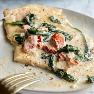 Creamy chicken breast topped with wilted spinach and sun-dried tomatoes on a white plate, with a gold fork beside the food.