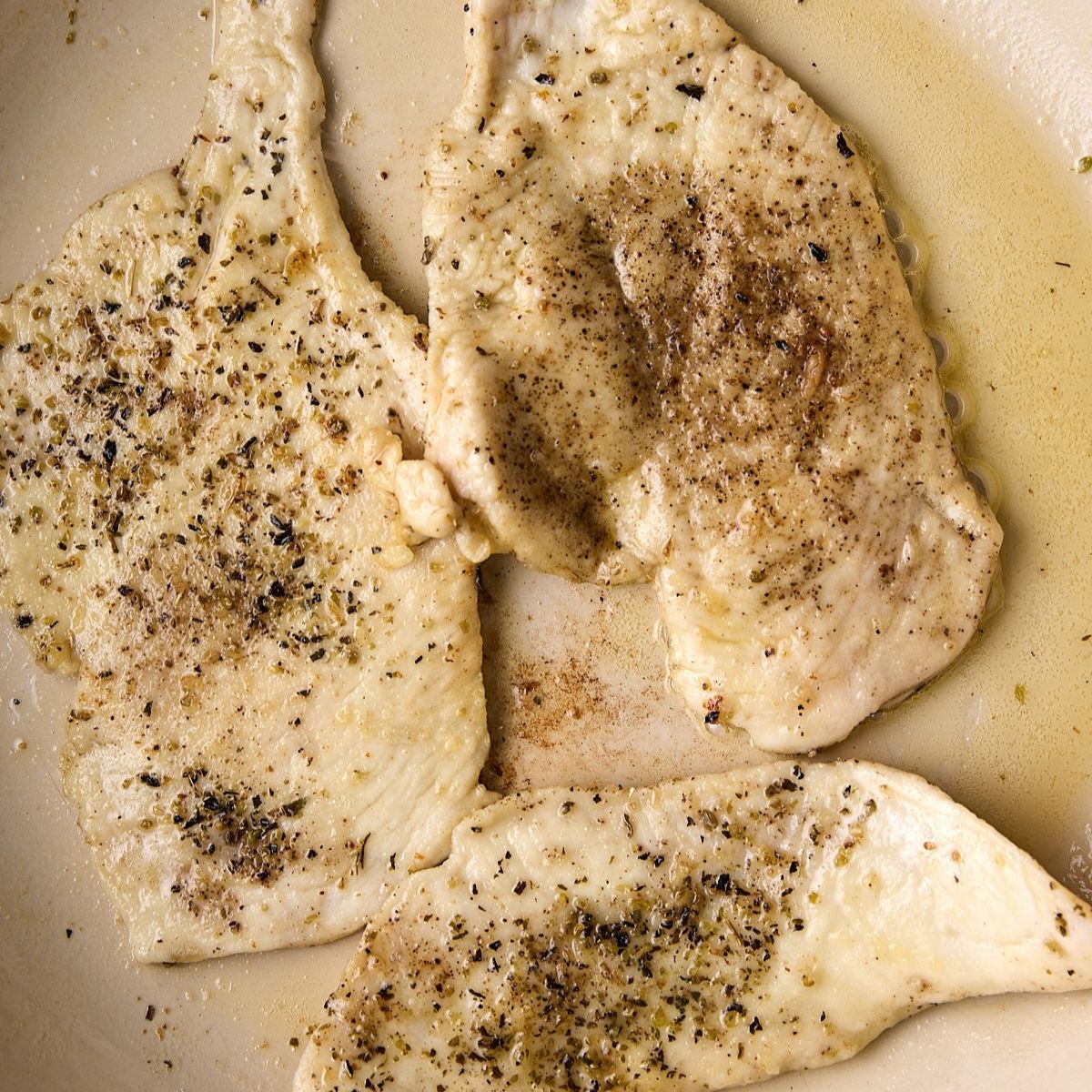 Chicken cutlets searing in a skillet, edges turning golden, flecked with black pepper and dried herbs.